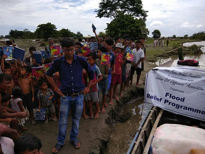 Children holding notebooks beside a flood relief programme banner near a boat